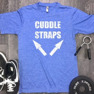 以下が含まれることがあります： 白い文字で「CUDDLE STRAPS」と書かれた青いTシャツと、2つの白い矢印。黒いスニーカーとグリップ強化剤も表示されています。5ポンドと2.3キロのウェイトプレートも表示されています。