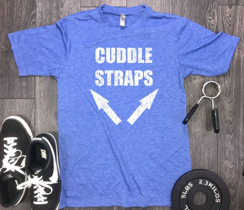 以下が含まれることがあります： 白い文字で「CUDDLE STRAPS」と書かれた青いTシャツと、2つの白い矢印。黒いスニーカーとグリップ強化剤も表示されています。5ポンドと2.3キロのウェイトプレートも表示されています。