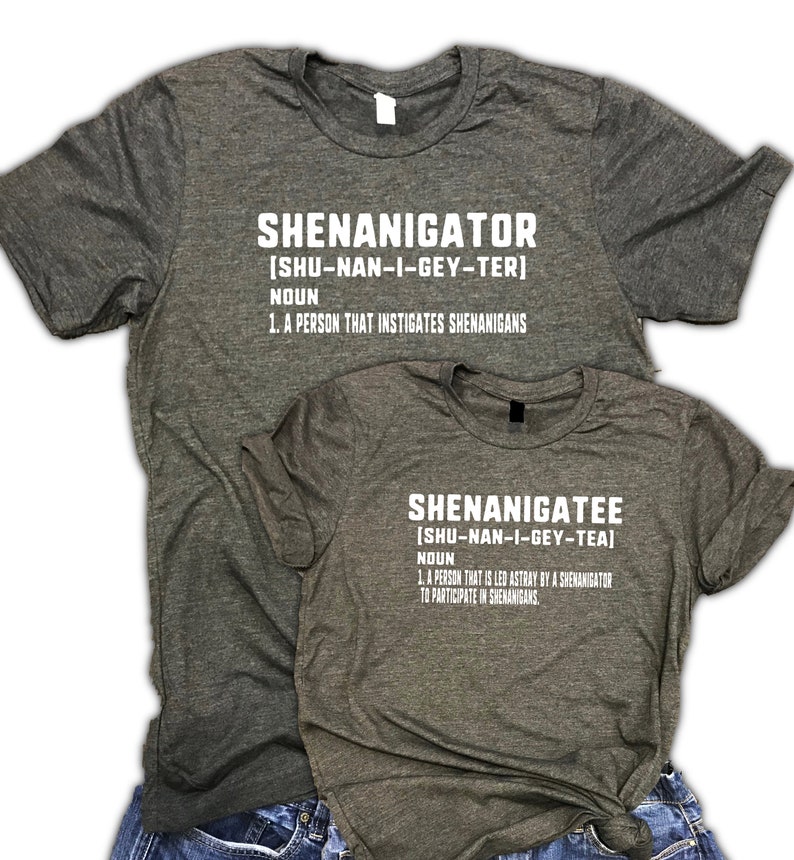 May include: Two heather gray t-shirts with white text. The top shirt says "Shenanigator [SHU-NAN-I-GEY-TER] NOUN 1. A PERSON THAT INSTIGATES SHENANIGANS". The bottom shirt says "Shenanigatee [SHU-NAN-I-GEY-TEA] NOUN 1. A PERSON THAT IS LED ASTRAY BY A SHENANIGATOR TO PARTICIPATE IN SHENANIGANS."