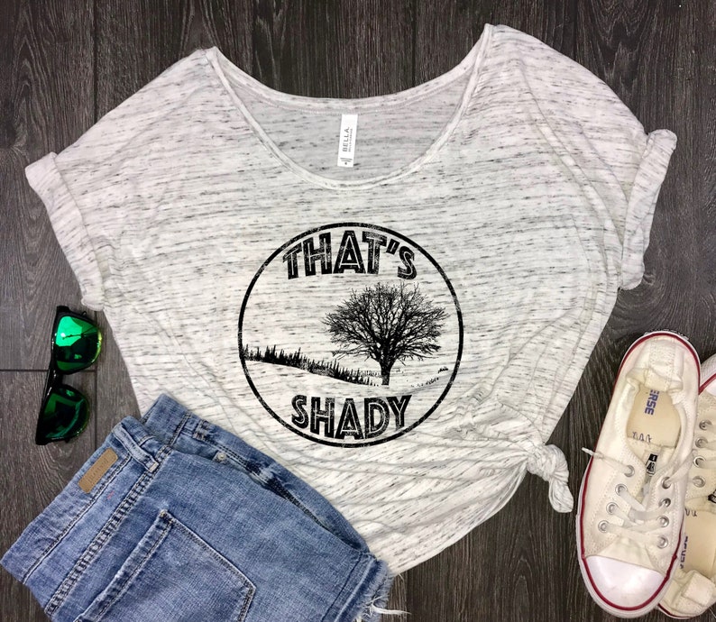 May include: A white and grey t-shirt with a black circle graphic that says "That's Shady" with a tree inside the circle. The shirt is on a wood background with a pair of blue denim shorts, a pair of white trainers, and a pair of green sunglasses.