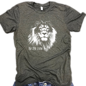 May include: A dark gray t-shirt with a white graphic of a lion's face and the text "BE THE LION".