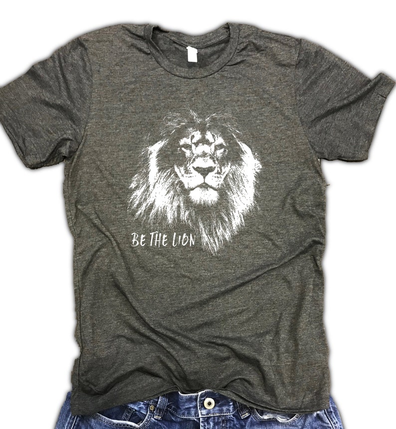 May include: A dark gray t-shirt with a white graphic of a lion's face and the text "BE THE LION".