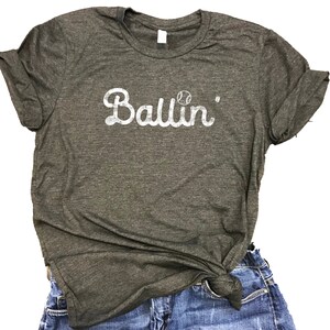 May include: A dark green t-shirt with the text "Ballin'" in white with a baseball graphic.