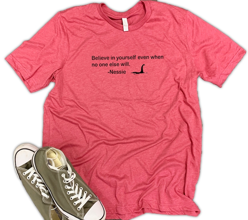 May include: A pink heather t-shirt with the text "Believe in yourself even when no one else will. -Nessie" and a black silhouette of the Loch Ness Monster.