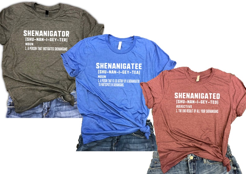 May include: Three t-shirts in different colours and designs. The first shirt is dark grey with the text "SHENANIGATOR [SHU-NAN-I-GEY-TER] NOUN 1. A PERSON THAT INSTIGATES SHENANIGANS". The second shirt is blue with the text "SHENANIGATEE [SHU-NAN-I-GEY-TEA] NOUN 1. A PERSON THAT IS LED ASTRAY BY A SHENANIGATOR TO PARTICIPATE IN SHENANIGANS". The third shirt is red with the text "SHENANIGATED [SHU-NAN-I-GEY-TED] ADJECTIVE 1. THE END RESULT OF ALL YOUR SHENANIGANS".