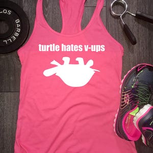 May include: A pink racerback tank top with the text "turtle hates v-ups" and a white turtle graphic. The tank top is next to a weight plate, a pair of sneakers, and a weight lifting grip.