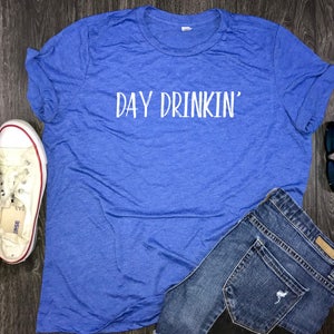 May include: A blue t-shirt with the text "DAY DRINKIN'" in white lettering. The shirt is laying on a wooden surface with a pair of white sneakers, blue jeans, and sunglasses.