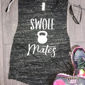 May include: Black and white muscle tank top with the text "Swole Mates" and a white kettlebell graphic. The tank top is on a wood surface with a pair of black and pink athletic shoes.