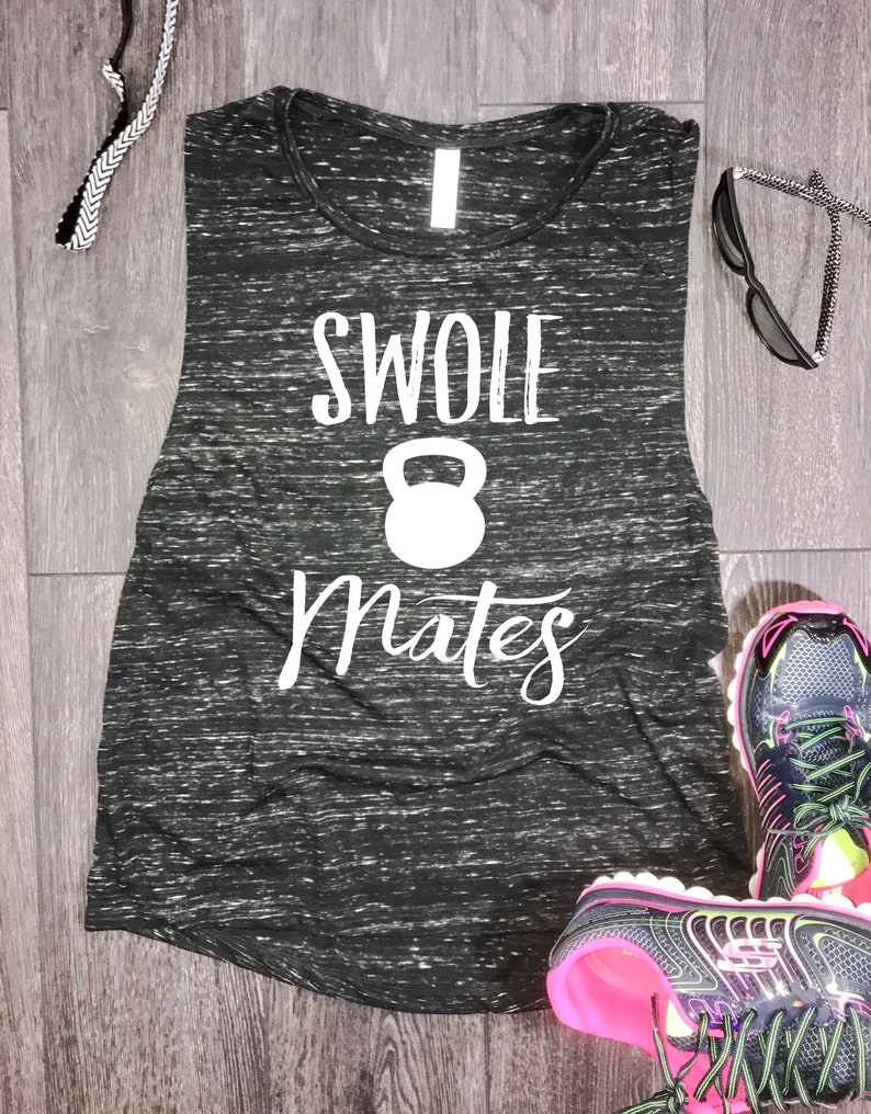 May include: Black and white muscle tank top with the text "Swole Mates" and a white kettlebell graphic. The tank top is on a wood surface with a pair of black and pink athletic shoes.