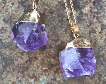 Könnte beinhalten: Zwei goldene Kettenhalsketten mit Amethyst-Kristallanhängern. Die Kristalle sind roh und haben einen violetten Farbton. Die Ketten sind zart und haben eine goldene Oberfläche.