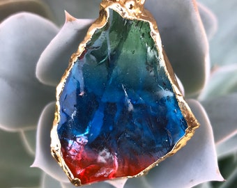 May include: A pendant featuring a multi-colored, iridescent glass stone with a gold-toned metal frame. The stone is a gradient of green, blue, and red.