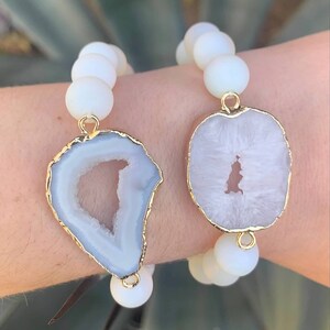 May include: Two white beaded bracelets with gold accents. Each bracelet features a large, white agate slice with a natural, open center. The agate slices are different shapes, one is a rounded oval and the other is a freeform shape.