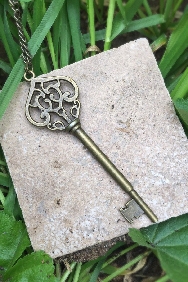 May include: A vintage-style key pendant necklace with a detailed ornate heart-shaped key design. The key is made of a brass-colored metal and hangs from a chain.
