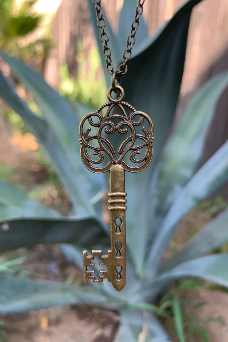May include: A gold-toned key pendant necklace with an ornate design. The key has a heart-shaped top and a detailed, intricate design.