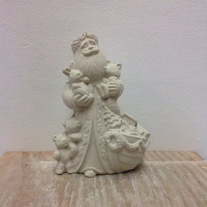 May include: A cream-colored Santa Claus figurine holding teddy bears and a basket of gifts. The Santa figure has a long white beard and is wearing a detailed robe. The figurine is on a wooden surface.