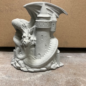 May include: A white ceramic sculpture of a dragon curled around a tower. The dragon has wings and a long tail. The tower has a pointed roof and windows.