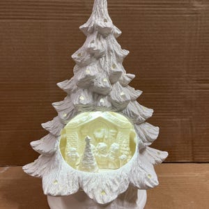 May include: A white ceramic Christmas tree with a detailed nativity scene inside. The tree has a textured, layered design and is illuminated with soft lighting. The base is a rounded shape, and the overall design is festive and decorative.