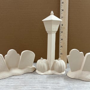 May include: Unpainted ceramic Halloween decorations. Includes a lamppost with pumpkins, a tombstone with the text "Cool Ghoul", and other tombstones. The decorations are unpainted and ready for customization.