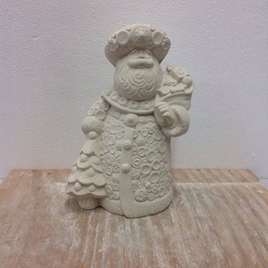 May include: A white ceramic figurine of a Santa Claus wearing a crown of flowers and a coat covered in buttons. He is holding a bag of gifts in one hand and a small Christmas tree in the other.