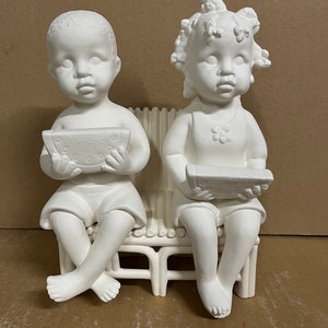 May include: Two ceramic figurines of children sitting on a bench. The boy is holding a slice of watermelon and the girl is holding a plate. Both figurines are painted white.
