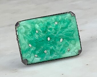 May include: A green jade brooch with a carved floral design, set in a silver tone metal frame.