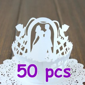 May include: 50 white cupcake cases featuring a cut-out design of a wedding couple standing under an archway. The cases are displayed on a white doily.