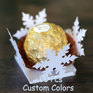 Könnte beinhalten: Fünfzig weiße, schneeflockenförmige Papierboxen mit einer goldenen Schokoladenbonbon darin. Die Boxen sind quadratisch angeordnet und der Text "50 Pcs Custom Colors" ist unter den Boxen gedruckt.