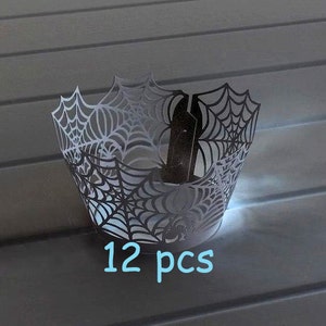 May include: Twelve black and white cupcake wrappers with a spiderweb design. The wrappers are made of paper and have a delicate, intricate design.