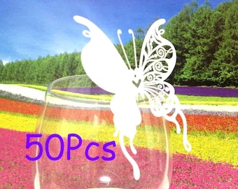 May include: 50 white paper butterfly place card holders. The butterflies are delicate and intricate with a lacy design. They are shown in a clear glass vase against a backdrop of colorful flowers.