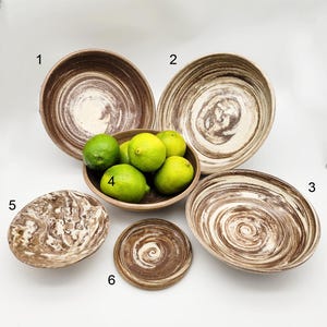 May include: A collection of handmade ceramic bowls and a small wooden bowl filled with limes. The bowls feature a brown and cream swirl pattern. The bowls vary in size and shape, including round and oval designs.
