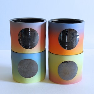 May include: Four ceramic cups with ombre glaze in shades of blue, orange, yellow, and pink. Each cup has a circular cutout revealing a black interior.