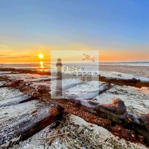 May include: A weathered wooden structure on a sandy beach at sunset. The sun is setting behind a lighthouse in the distance. The sky is a mix of orange, pink, and blue.