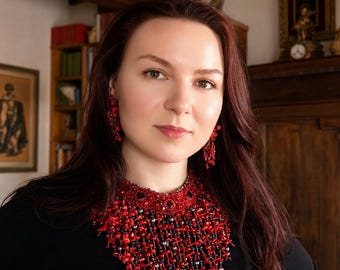 May include: A statement necklace and matching drop earrings featuring a dense arrangement of small, irregular red coral-like beads interspersed with tiny dark metallic accents, worn against a plain black top to highlight the vibrant red color.
