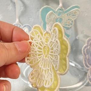 May include: Close-up of a yellow and white angel ornament with intricate lace-like detailing. The angel has a yellow body and white wings, with a ribbon for hanging. Other angel ornaments in blue and purple are visible in the background.