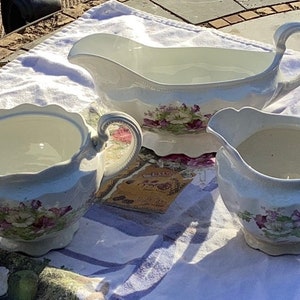 May include: A three-piece porcelain serving set. The set includes a gravy boat, a creamer, and a sugar bowl. Each piece is white with a floral pattern in shades of purple, pink, and green. The set is placed on a white cloth.