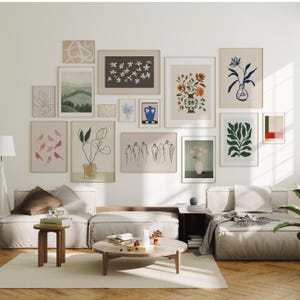 May include: A living room with a white wall featuring a gallery wall of framed art prints. The art prints feature various designs, including floral patterns, abstract shapes, and nature scenes. The room is furnished with a light-colored sectional sofa, a coffee table, and a side table.