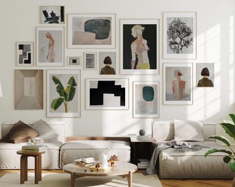 May include: A living room interior with a white wall featuring a gallery wall of framed artwork. The artwork includes abstract paintings, black and white geometric designs, and illustrations of people and plants. The room is furnished with a light grey sectional sofa, a wooden coffee table, and a side table with a lamp.
