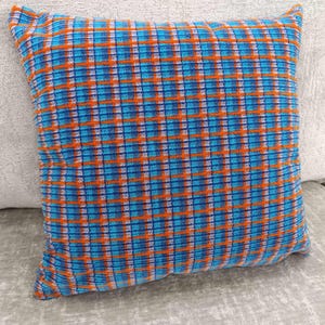 May include: Square cushion featuring a colourful plaid design in blue, orange, and white. The cushion is made from a textured fabric and is placed on a light grey surface. The cushion has a classic, decorative design.