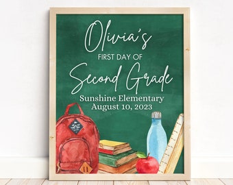 May include: A green chalkboard with the text "Olivia's First Day of Second Grade" and the date "August 10, 2023". The chalkboard is decorated with a red backpack, a stack of books, a red apple, a blue water bottle, and a wooden ruler.