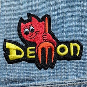 May include: An embroidered patch featuring a red cartoon character with large white eyes holding an orange pitchfork. The word DEMON is stitched in bright yellow letters across the bottom of the black background, which is attached to blue denim fabric.