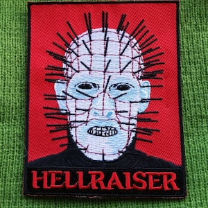 May include: A red and black embroidered patch featuring a character with a grid-like face and many spikes. The text "HELLRAISER" is written in red letters on a black background.
