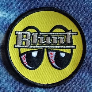 May include: Embroidered patch with a yellow background, black border, and the word "Blunt" in silver and gray. The patch features cartoon eyes with red veins, giving a relaxed vibe. The patch is round and likely for clothing or accessories.