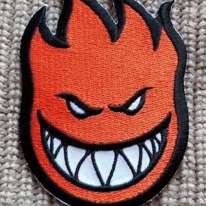 May include: Orange and black embroidered patch with a flame design and a grinning face with sharp teeth.