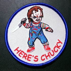 May include: A white embroidered patch with a blue border. The patch features a cartoon image of a red-haired doll holding a knife and the text "Here's Chucky".