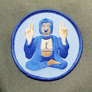 May include: A blue embroidered patch with a light blue background featuring a seated figure with a peaceful expression. The figure has a blue robe and is in a meditative pose with hands raised in a gesture of peace.