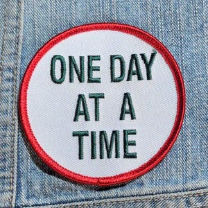 May include: A circular embroidered patch with a white background and a bold red border. The text "ONE DAY AT A TIME" is stitched in dark green capital letters across the center of the patch, which is placed on a blue denim fabric surface.