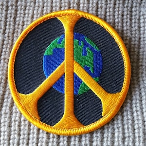 May include: A yellow peace sign with a blue and green globe in the center. The peace sign is embroidered on a black fabric patch.