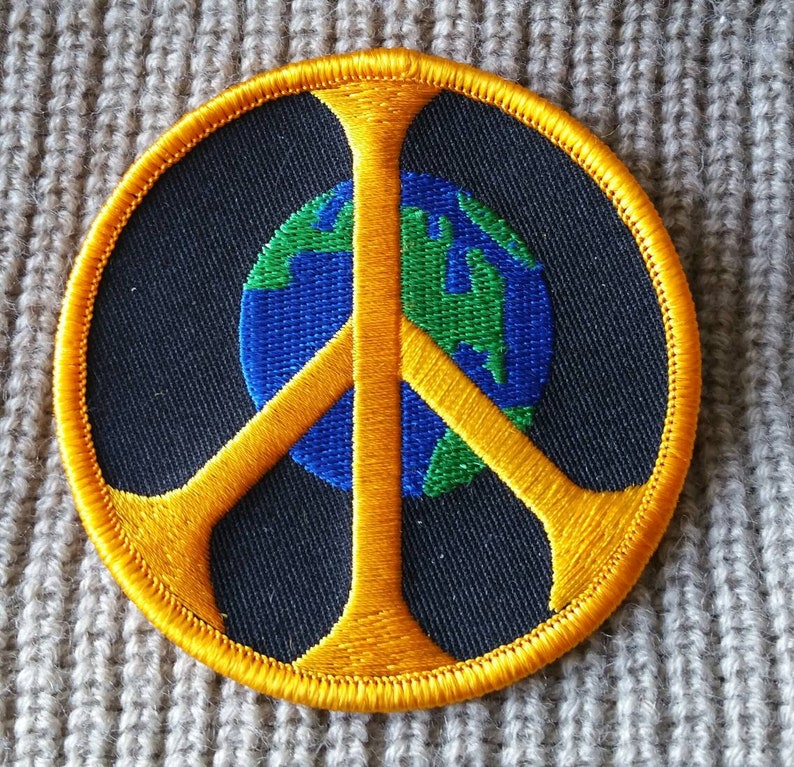 May include: A yellow peace sign with a blue and green globe in the center. The peace sign is embroidered on a black fabric patch.