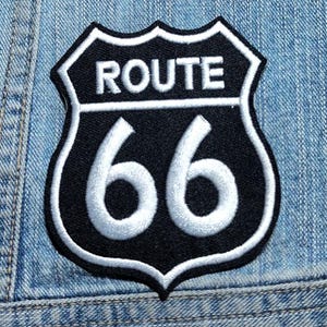 May include: A shield-shaped embroidered patch featuring a black background with a white border. The text ROUTE is embroidered in white at the top, and the number 66 is embroidered in large white numerals below a horizontal white line.