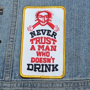 May include: A rectangular embroidered patch with a white background and a yellow border. It features a red line-art illustration of a person with a mustache above the text NEVER TRUST A MAN WHO DOESN'T DRINK in black and red stitched lettering.